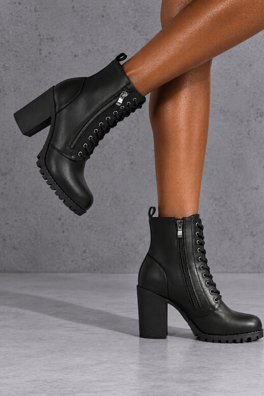 A pair of black lace-up ankle boots with chunky block heels and side zippers, shown on a model's feet against a gray concrete-textured background.