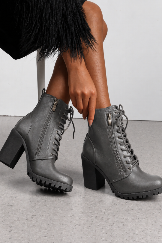 A pair of dark gray faux-leather lace-up ankle boots with block heels, side zippers, and lug soles, shown on a model's feet against a light gray floor and white wall.