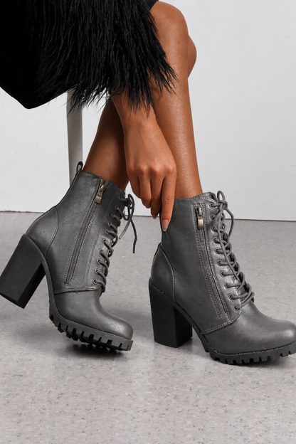 A pair of dark gray faux-leather lace-up ankle boots with block heels, side zippers, and lug soles, shown on a model's feet against a light gray floor and white wall.