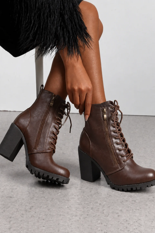 A pair of dark brown faux-leather lace-up ankle boots with chunky black block heels, shown on a model sitting on a chair. The boots feature side zippers and rugged lug soles.