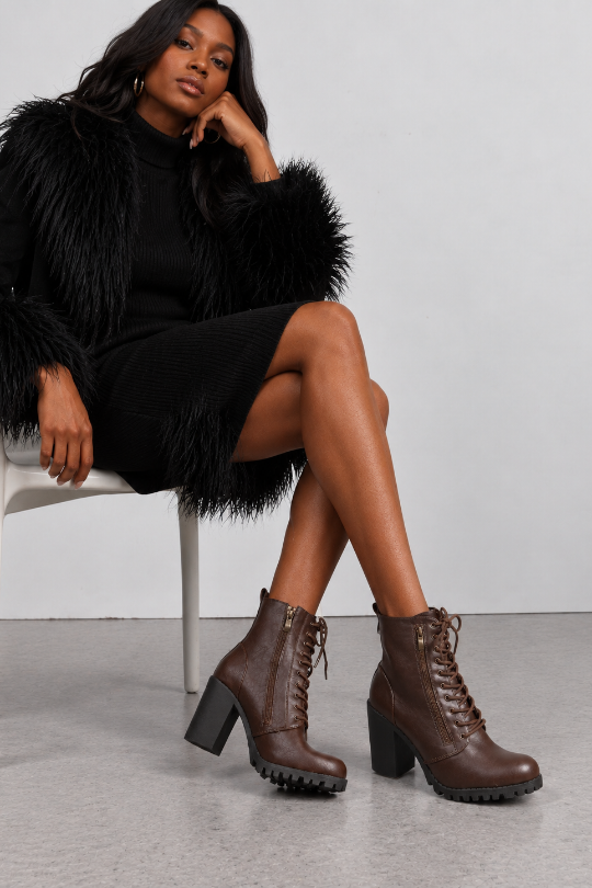 A woman seated on a white chair wearing dark brown lace-up ankle boots with chunky block heels, posed in a studio with a light gray background.