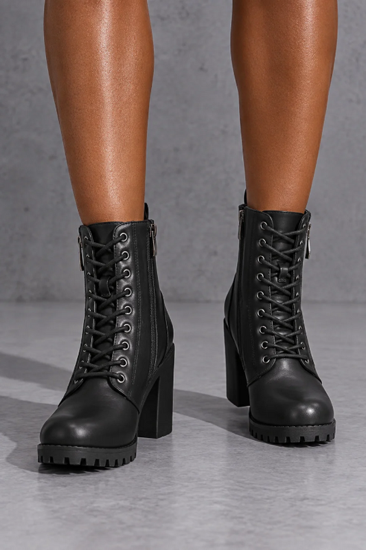 A pair of black lace-up ankle boots with block heels and lug soles, shown on a model's feet against a gray concrete floor and wall.